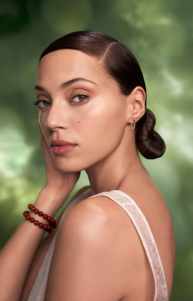 A woman with slicked-back dark hair in a low bun, styled at an Aveda salon, poses with one hand on her face, wearing a sleeveless white top, red bead bracelet, and small hoop earring against a green background. - Studio A Salon Spa in Osceola, WI