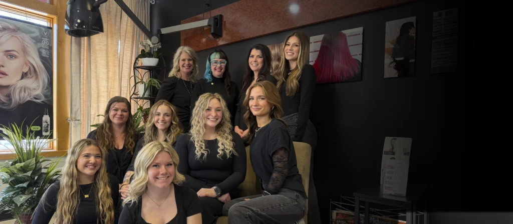 A group of eleven women, all dressed in black, gather in a well-lit Aveda salon in Osceola, Wisconsin, surrounded by plants and hair color posters on the walls. - Studio A Salon Spa in Osceola, WI
